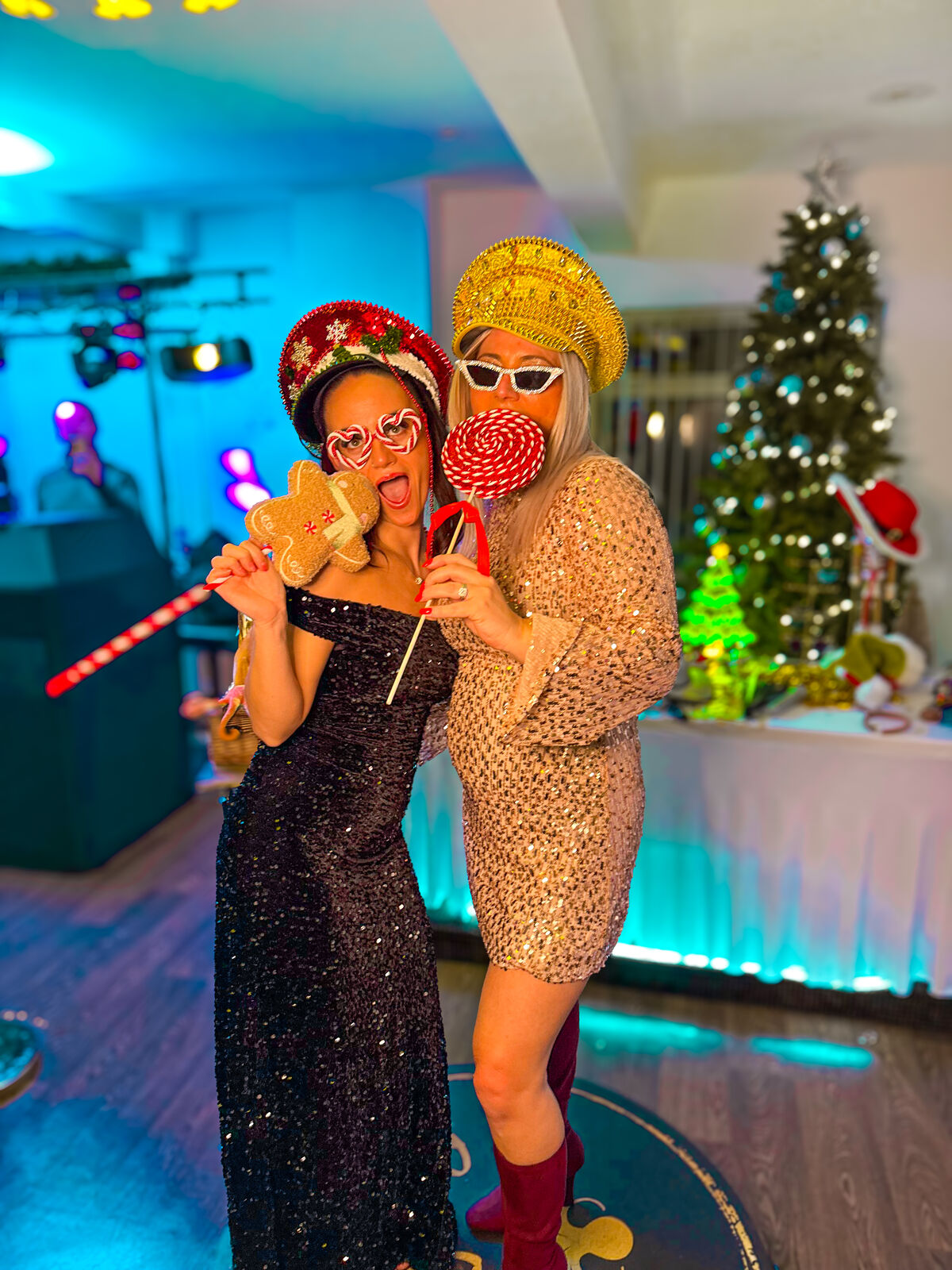 Two guests in sequin dresses with festive hats, candy-cane glasses and giant lollipop props at a Christmas party